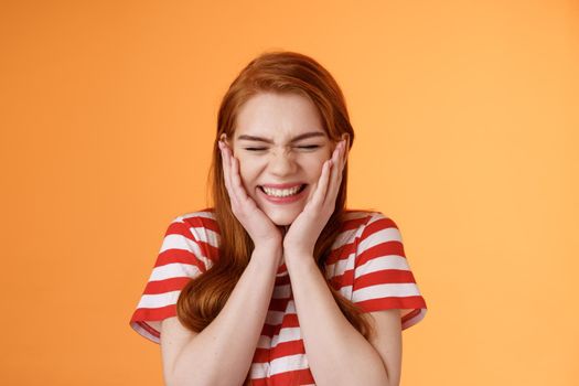 Close-up cheerful lucky excited smiling girl, cannot believe cheering hear excellent news, close eyes celebrating, touch cheeks blushing joy delight, rejoicing awesome situation, orange background.