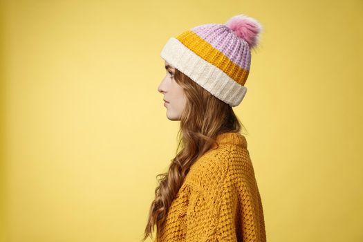 Profile shot attractive female curly hairstyle wearing hat knitted sweater standing queue order hot warm drink look left normal casual expression, posing yellow background studio. Copy space