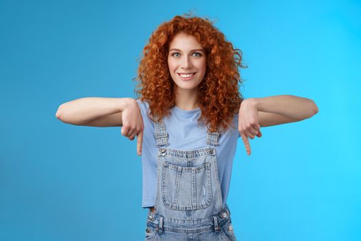 Lifestyle. Charismatic sassy flirty redhead daring ginger girl curly haircut pointing down index fingers smiling broadly enthusiastic explore new store pointing promo like cool advertisement blue background.