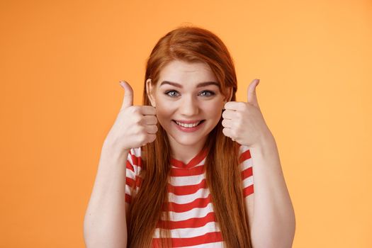 Hopeful cheerful cute redhead girlfriend rooting for you, believe win, smiling supportive satisfied, show thumbs-up delighted, approve choice, wish good luck, encourage friend, orange background.