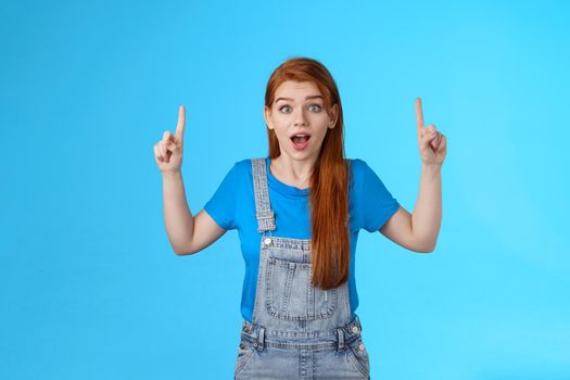 Excited cheerful cute ginger girl 20s, open mouth entertained, look enthusiastic amazed, pointing up, top copy space, promote excellent awesome new product, stand blue background astonished.