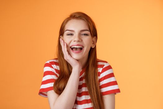 Cute cheerful smiling redhead woman talking friends, laughing out loud happily showing healthy toothy white smile, touch cheek silly, lovely giggling funny conversation, happy orange background.
