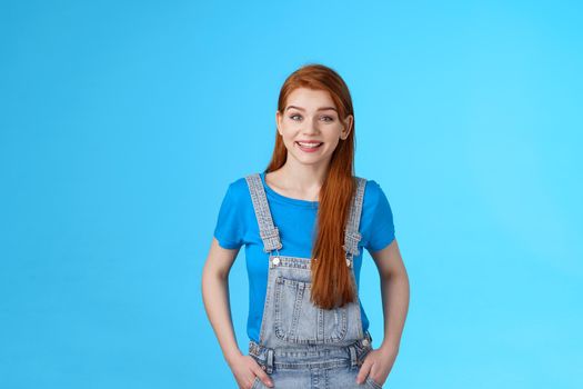 Cheerful glad redhead woman standing joyful relaxed pose, wear denim dungaress over t-shirt summer outfit, smiling pleased, carefree friendly conversation, pose blue background. Copy space