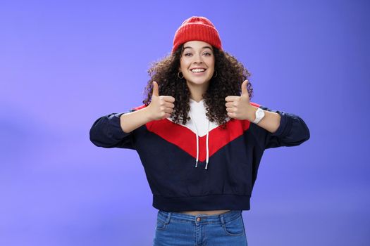 Girl totally agrees recommends cool place showing thumbs-up and smiling excited with happy grin standing upbeat and delighted against blue background in warm beanie and sweatshirt. Body language and emotions concept
