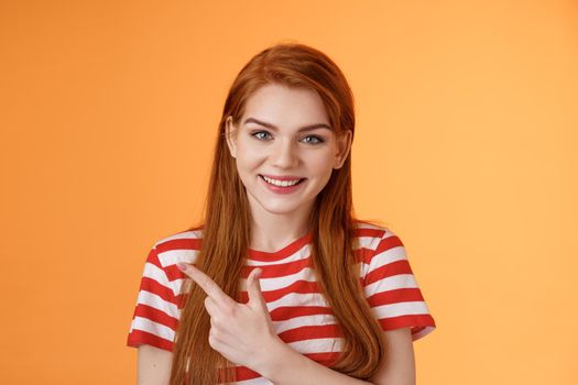 Close-up cheerful smiling girl pointing sideways. Redhead woman introduce promo gladly recommend good offer, grin discuss interesting place indicate upper left corner, orange background.