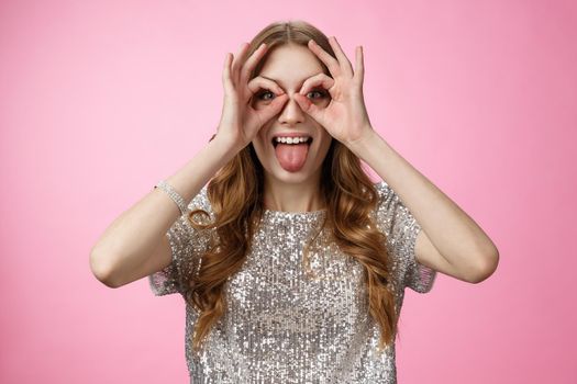 Carefree playful funny young party girl fool around showing tongue immature mimicking make goggles from hands widen eyes look through circles, having fun entertain friends, standing pink background.