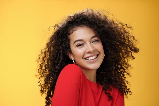 Romantic and flirty young cute caucasian girlfriend with curly hair in red blouse making silly pose smiling joyfully and lifting shoulder coquettish smiling broadly at camera over yellow wall. Love, relationship, seduction concept