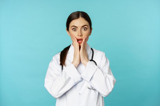 Portrait of shocked woman doctor, female hospital intern in white coat, looking concerned and confused at camera, disbelief face, standing over torquoise background.