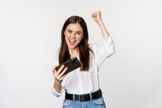 Happy brunette woman playing mobile video game, smiling and looking at screen excited, standing over white background.