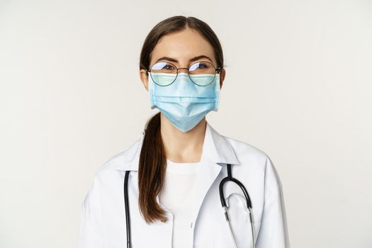 Portrait of medical worker, female physician in face mask from covid during pandemic, smiling and looking enthusiastic, standing over white background.