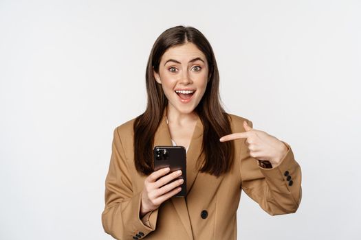 Enthusiastic saleswoman, business woman pointing finger at mobile phone and smiling, showing on cellphone, standing against white background.