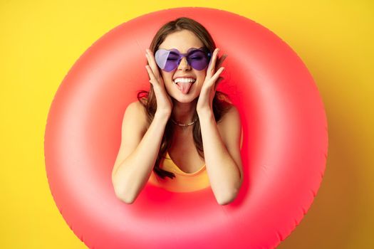 Close up portrait of enthusiastic young woman inside pink swimming ring, laughing and smiling, enjoying beach holiday, summer vacation, yellow background.