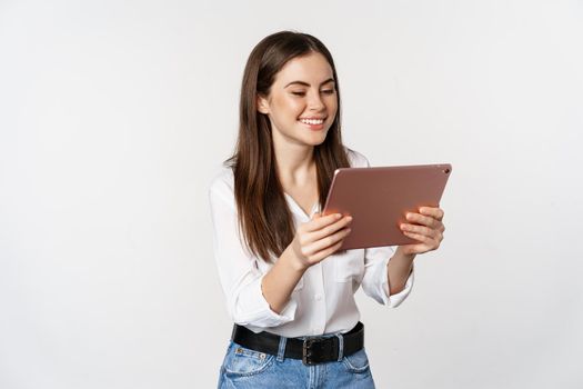 Portrait of beautiful girl smiling, looking happy, watching on digital tablet, standing against white background. Copy space