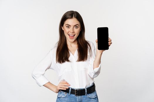 Happy smiling woman showing smartphone app interface, mobile phone screen, demonstrating promo offer, standing over white background.