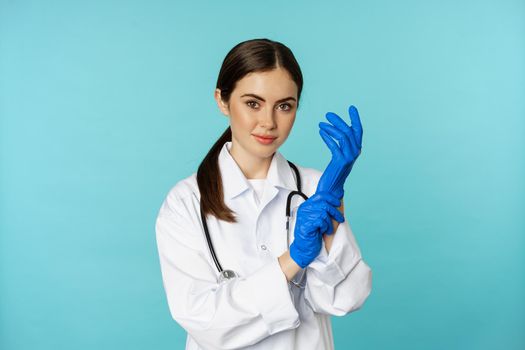 Young doctor, professional physician woman, put on gloves, smiling at camera, ready for medical checkup, standing in uniform over blue background.