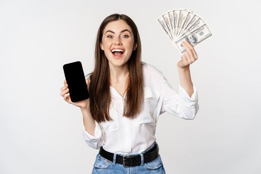 Enthusiastic young woman winning money, showing smartphone app interface and cash, microcredit, prize concept, standing over white background.