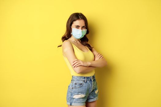 Covid-19 vaccination. Beautiful young woman in face medical mask got vaccinated during pandemic, standing over yellow background.