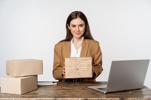 Businesswoman, store owner sitting near laptop, holding box with client order and smiling, working on computer, white background.