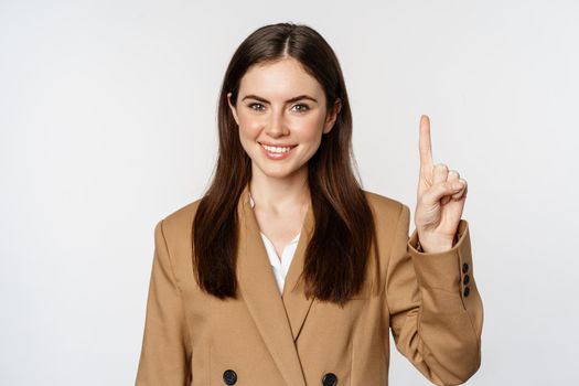 Portrait of corporate woman, saleswoman showing number one finger and smiling, standing in suit over white background.