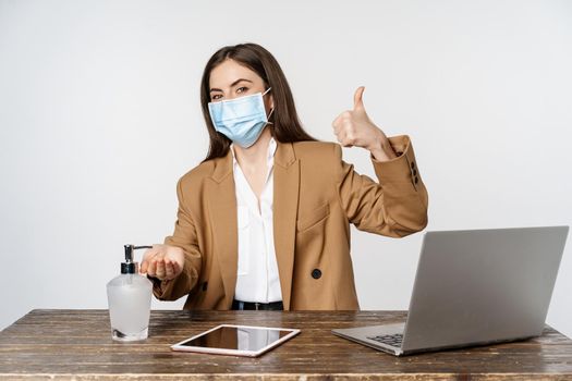 Workplace and covid-19 concept. Smiling businesswoman in face medical mask, showing thumbs up while using hand sanitizer cleanser to clean wash hands, white background.