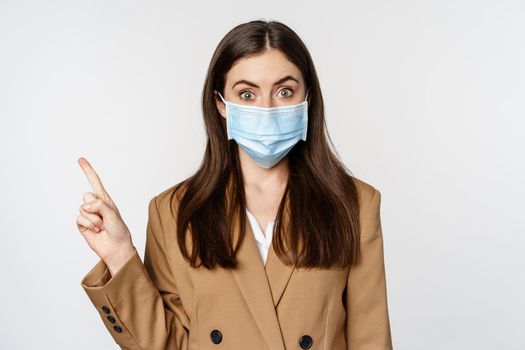 Coronavirus and people concept. Portrait of business woman at workplace wearing face mask, pointing finger left at logo, company banner, white background.