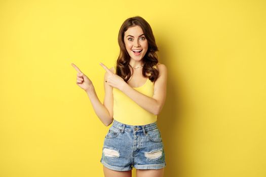 Portrait of stylish summer girl, brunette woman showing way, pointing fingers left, advertising, standing over yellow background. Copy space