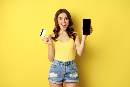 Enthusiastic modern woman showing smartphone screen, holding credit card and smiling, recommending mobile app, standing over yellow background.