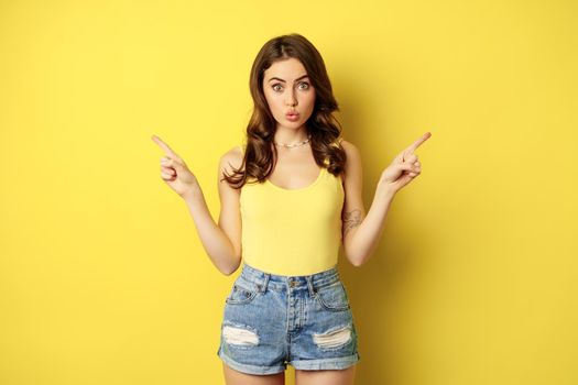 Portrait of stylish young female model, showing advertisement, pointing at logo or banner, demonstrating two ways, choices in store, standing over yellow background.