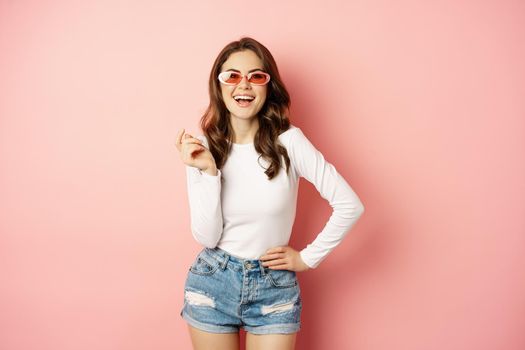 Portrait of stylish glam girl in sunglasses, laughing and smiling, standing over holiday pink background. Copy space