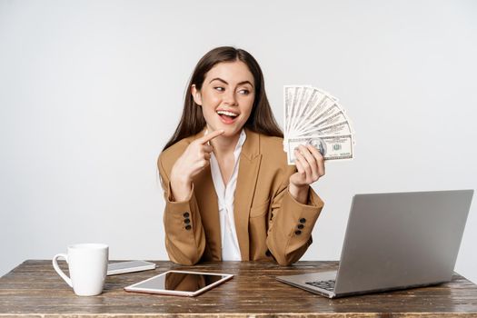 Portrait of businesswoman sitting in office with money, working and making profit income, posing happy against white studio background.