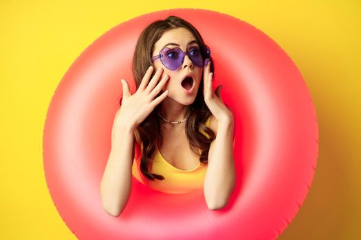 Portrait of young pretty woman in sunglasses, wearing beach swimming ring, looking surprised and amazed, shocked wow face, yellow background.