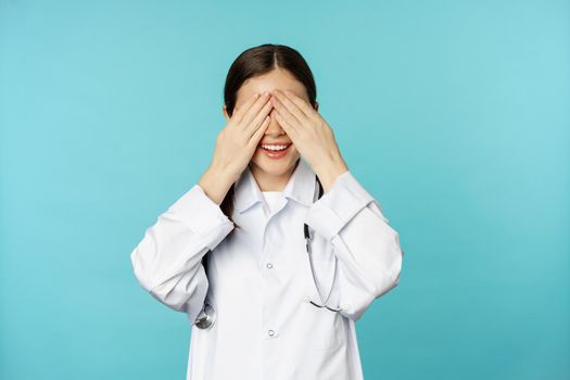 Smiling happy therapist, woman doctor cover close eyes with hands and waiting for surprised, anticipating, standing in white coat over torquoise background.