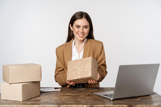 Businesswoman, store owner sitting near laptop, holding box with client order and smiling, working on computer, white background.