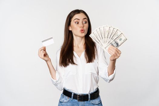 Excited woman holding credit card and money, looking amazed at cash, standing against white background. Copy space