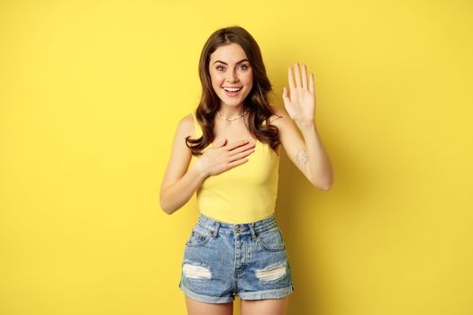 Enthusiastic girl raising hand, promise, introduce herself, say her name, standing over yellow background, smiling and looking excited.