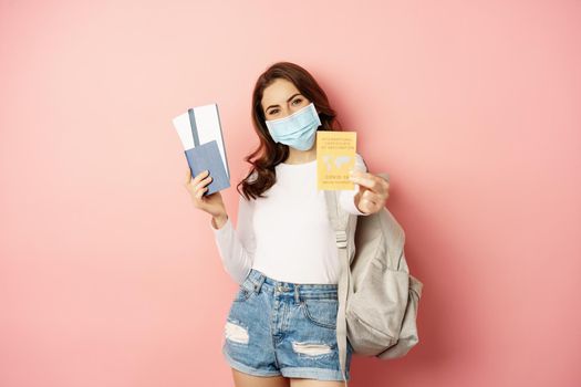 Young woman going on trip, wearing medical mask, showing covid certificate, passport and aiplance tickets, vacation during pandemic, pink background.