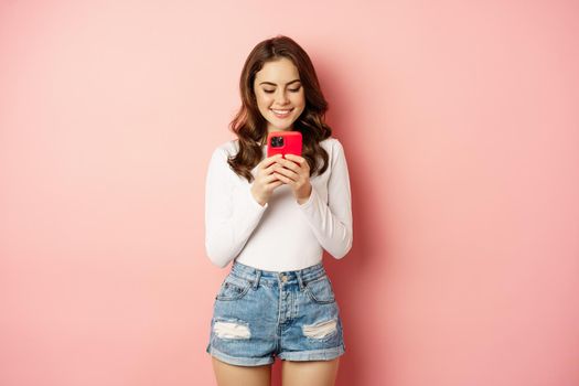 Smiling brunette girl using smartphone, texting message, order online on mobile phone app, standing over pink background. Copy space
