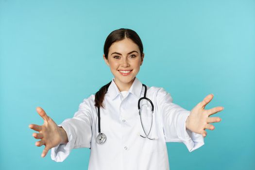 Smiling friendly doctor, girl healthcare worker, intern reaching hands, inviting, hugging or receiving in arms, standing over torquoise background.