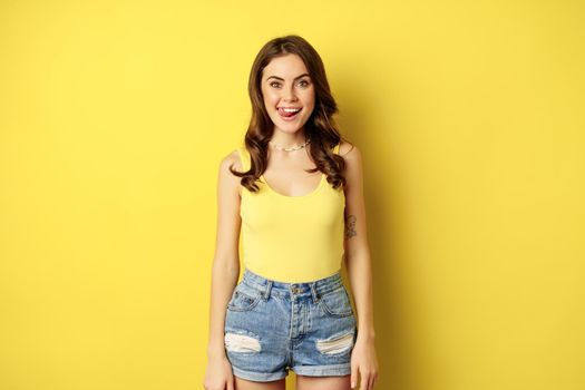 Stylish young brunette woman showing her white healthy teeth, smiling and showing tongue, winking enthusiastic, standing over yellow background.