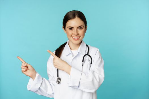 Portrait of smiling medical worker, girl doctor in white coat with stethoscope, pointing fingers left, showing medical clinic advertisement, torquoise background.