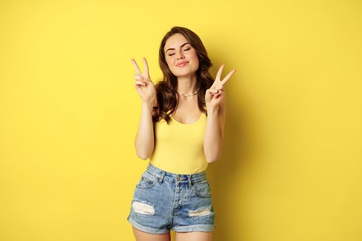 Positive girl, female model showing peace, v-sign gesture and smiling, standing in tank top and denim shorts, yellow background.