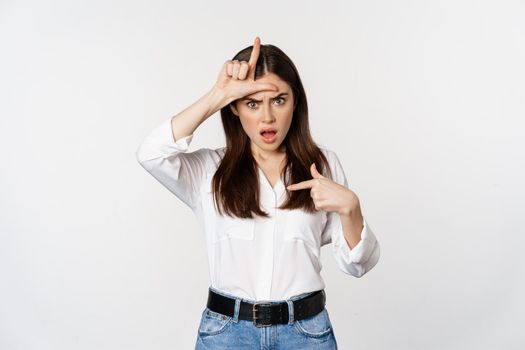 Insulted woman showing loser L word sign on forehead, pointing at herself frustrated, standing over white background.