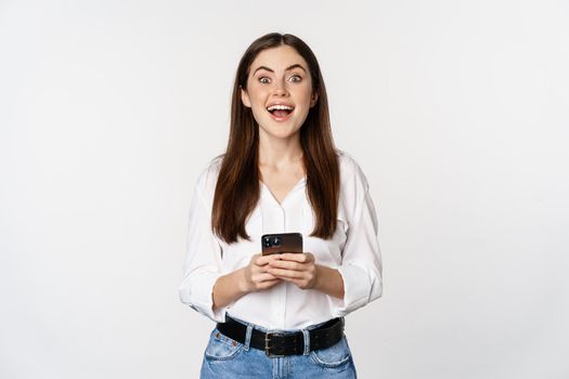 Young woman with smartphone, smiling and looking at camera, using mobile phone app, cellular technology and online shopping concept, white background.