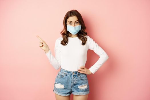 Beautiful stylish woman in medical face mask from covid, pointing finger at upper left corner, showing advertisement, standing over pink background.