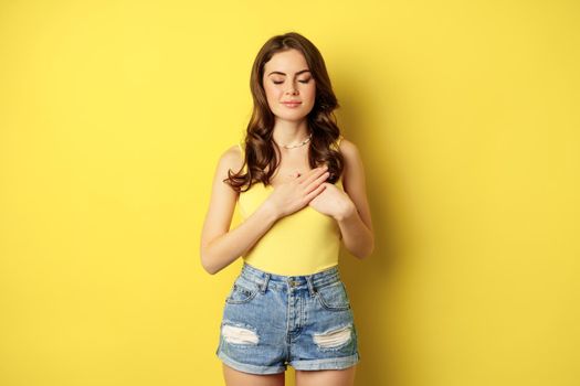 Young woman in tank top, holding hands on heart with care and tenderness, smiling at camera, looking grateful, thankful, standing over yellow background.