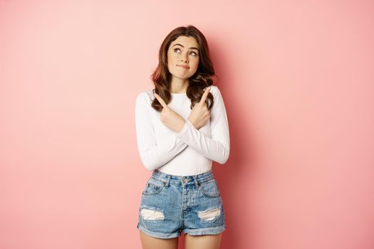 Complicated woman looking indecisive, stylish girl pointing fingers sideways and gazing thoughtful at upper left corner, making choice, deciding, pink background.