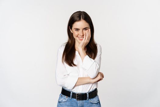 Beautiful young brunette woman, gently touches her face skin and smiling coquettish, tender pose against studio white background.