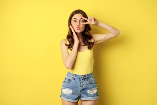 Beautiful glamour girl in summer outfit, showing peace sign, kissing face, standing against yellow background.