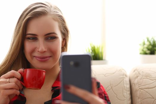 Young beautiful woman drinks coffee and looks into smartphone. Positive online communication concept