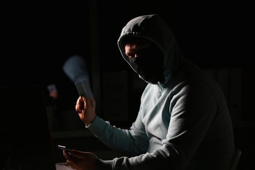 Hacker in dark jacket looking at laptop screen in dark room is holding bank card. Network security and protection concept